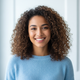Smiling young woman with curly brown hair, wearing a soft sky blue sweater, against a bright white background.